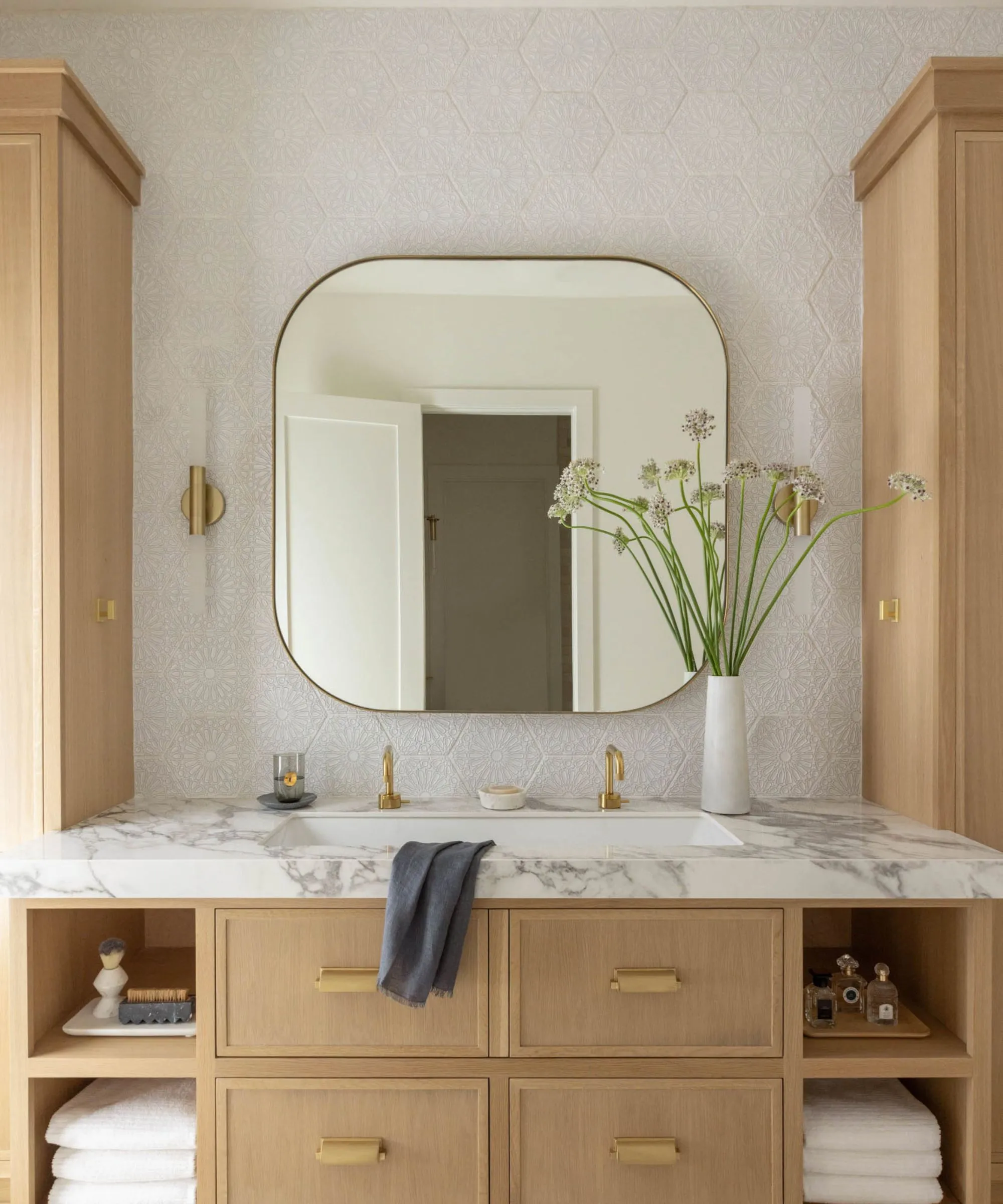 אזור אמבטיה ניטרלי עם אגרטל מראה מרובע מעוגל ופרחים משטח שיש מגבת קטנה וכיור לבן עם ברזי זהב. מתחת מגירות חומות ומדפים.' loading='lazy' title=