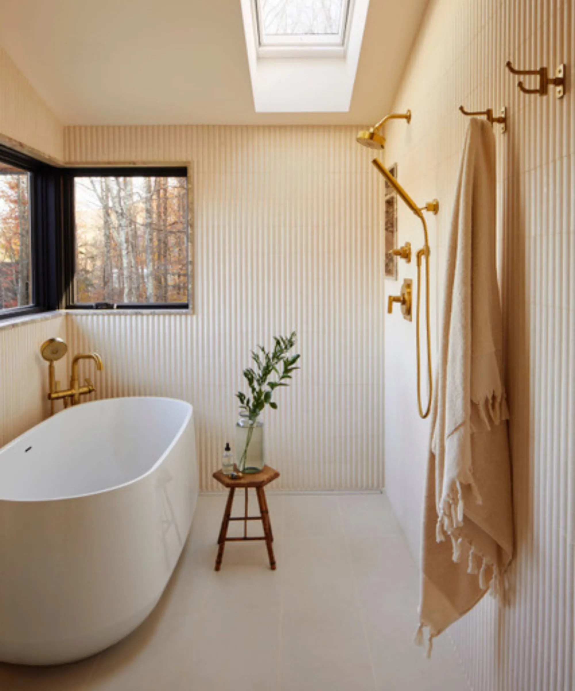 חדר רטוב עם קירות מפוספסים אמבטיה בודדת, מקלחת זהב ומגבת וו ושרפרף עץ' loading='lazy' title=