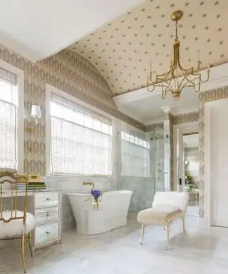 Grande salle de bains luxueuse avec plafond et murs recouverts de papier peint à motifs crème, baignoire blanche, douche en verre, siège, sol et carrelage mural en marbre, lustre doré,