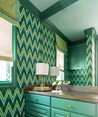 Salle de bain verte avec papier peint vert en zigzag, plafond peint en vert et garniture de plafond vert foncé, armoire verte avec dessus en pierre marron foncé, lampe de table avec abat-jour blanc