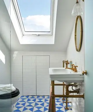 Salle de bains d'un blanc éclatant avec murs et plafond peints en blanc, carrelage géométrique bleu et blanc, puits de lumière sur plafond incliné, lavabo blanc traditionnel avec tuyauterie métallique apparente, miroir arrondi en métal et applique murale en métal et verre