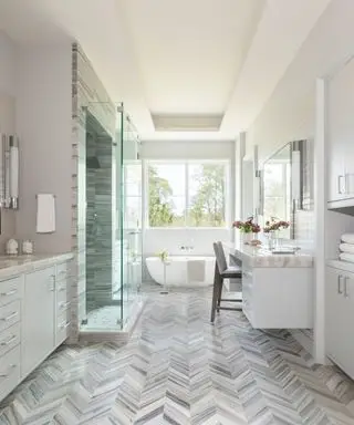 Grande salle de bain avec plafond blanc encastré, sol en carrelage gris chevrons finition marbre, donnant sur petite baignoire blanche, douche avec porte vitrée, armoires blanc cassé avec comptoir en marbre, coin salon, murs carrelés