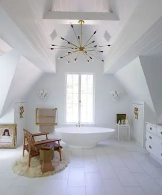 Une grande salle de bain avec une baignoire blanche sous une fenêtre, un fauteuil en bois et du carrelage blanc au sol