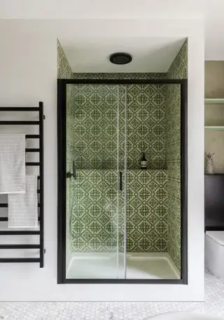 Ducha con azulejos con motivos verdes y estante, puertas de cristal con marco negro y radiador colgado en la pared en color negro.