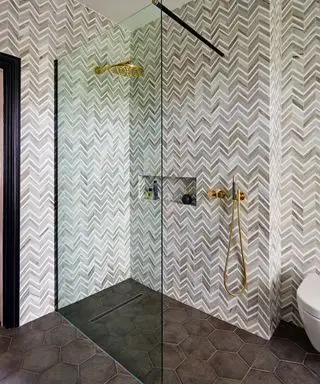 Sprchový kout ilustrovaný šedými dlaždicemi rybí kosti a zlatými akcenty.