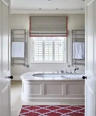 Une grande baignoire au centre d'une salle de bains blanc cassé avec un tapis de sol rouge et blanc