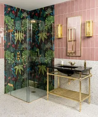 Un esempio di idee per una mensola da bagno che mostra un bagno piastrellato rosa con cabina doccia rivestita con carta da parati a motivi tropicali e un mobile lavabo dorato aperto