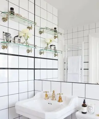 Un esempio di idee per mensole da bagno che mostrano un bagno piastrellato bianco con due ripiani in vetro montati a parete accanto a un lavandino bianco