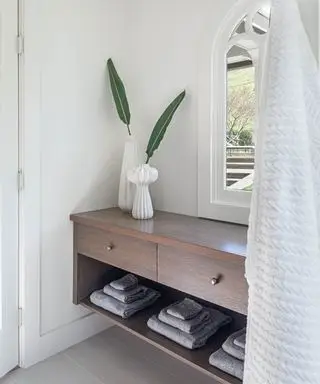 Bagno dallo stile rilassato e minimalista con scaffali in legno scuro, contenitori aperti e chiusi, ripiani bassi aperti in cui riporre asciugamani bianchi, due vasi decorativi con steli di foglie, finestra ad arco, pareti dipinte di bianco.