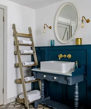 Bagno tradizionale in stile country con pavimento piastrellato, pannelli verniciati blu e coordinati, lavabo in legno, specchio rotondo con cornice color crema, due applique metalliche su entrambi i lati, scala in legno che funge da scaffale, asciugamani riposti su ogni sezione