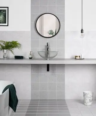 Une salle de bain carrelée grise avec comptoir, miroir rond et lavabo en verre transparent.