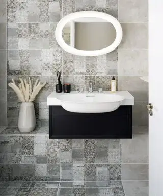 Salle de bains carrelée grise avec effet mosaïque unique, lavabo noir et blanc, miroir blanc arrondi et vase décoratif avec herbes