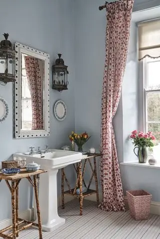 Un exemple d'idées de salle de bain bleue montrant une salle de bain bleu pastel dans un décor d'époque, des rideaux imprimés roses, un miroir orné, des appliques murales et des éléments latéraux en rotin.
