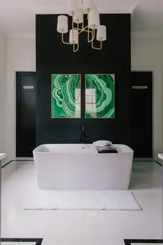Une baignoire rectangulaire blanche dans une salle de bains avec des murs noirs et du carrelage blanc, des œuvres d'art vertes modernes sur le mur et une suspension dorée et blanche