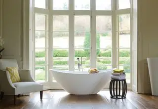 Une baignoire blanche dans une grande fenêtre avec un fauteuil et un tabouret en bois avec des serviettes dessus