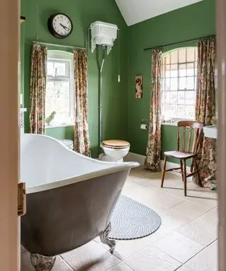 Baño pintado de verde con cortinas florales y bañera independiente pintada de gris