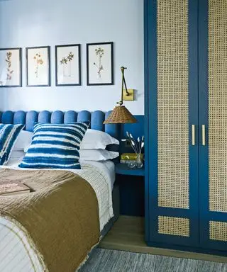 Blekblåmalt soverom, hodegjerde i indigo, garderobe med rottingdører i matchende indigo nyanse, hvitt sengetøy med hvite og indigo stripete puter, brune plagg, matchende kunstverk over sengen som viser pressede blomster, liten nattbordshylle, vegglampe med vevd fletteskjerm ovenfor.