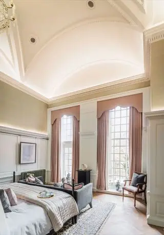 Veľká architektonická krémová spálňa so sivou pohovkou, ružovými závesmi a kreslom