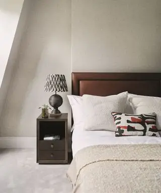 Un lit rembourré aux sourcils foncés avec une literie blanche et un jeté beige. Une table de chevet noire avec une lampe noire