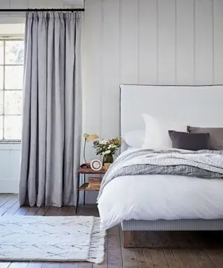 En lysegrå soveromsgardin med blendingsfôr i et soverom med hvit seng og hvite veggpaneler