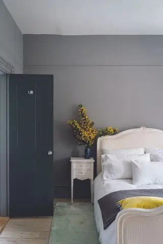 chambre grise avec tapis vert pâle, lit blanc à la française, table d'appoint assortie, fleurs jaunes, coussin jaune, porte peinte en gris foncé