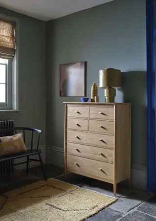 grønt soverom med tallboy, steingulv, teppe, lampe, sokler, kunstverk, ercol benk og pute, persienner ved vinduet, elektrisk blå dørkarm