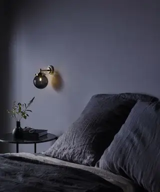 Un exemple de planification de l'éclairage d'une chambre montrant un mur gris foncé avec une applique murale sphérique noire derrière un lit gris foncé.