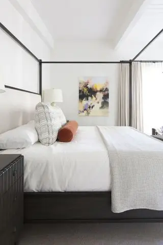Dormitorio blanco de Melinda Mandell con cama negra, cojín naranja y obras de arte