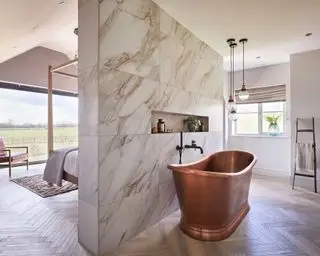 욕조가 있는 넓고 현대적인 침실, 대리석 칸막이 벽, 구리 욕조, 나무 바닥, 매달린 펜던트 조명 세 개, 침대, 가죽, 배경의 나무 안락의자, 천장부터 바닥까지 내려오는 대형 창문으로 구분되어 있습니다.