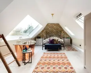 아치형 천장, 채광창 2개, 침대 뒤의 벽지 벽, 금속 침대 프레임, 패턴 러그 2개, 채광창 옆 구리 욕조가 있는 로프트 스타일 침실,