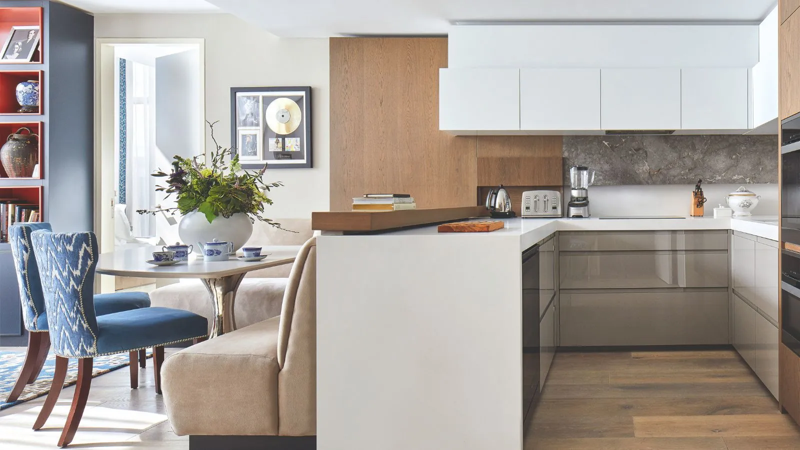 Une cuisine lumineuse avec armoires blanches et parquet. À gauche se trouve un coin salon/salle à manger confortable avec un canapé-lit beige et des sièges à motifs bleus avec des pieds en bois. À l'extrême gauche se trouve une étagère bleue.' loading='lazy' title=