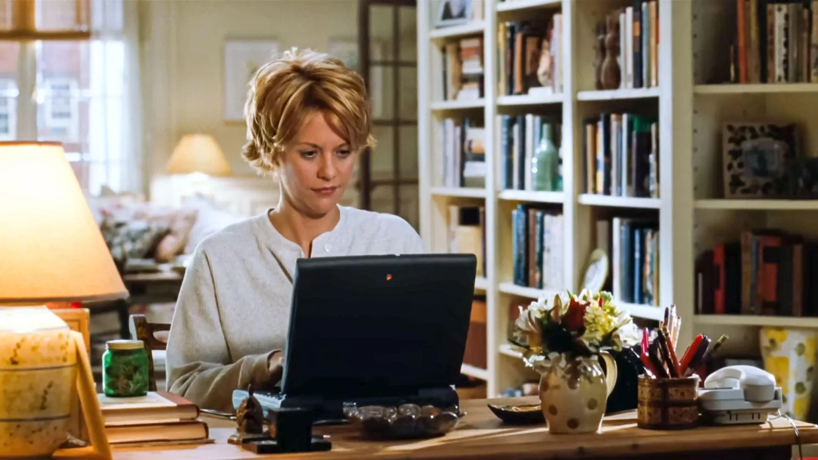Meg Ryan dans You Got Got Mail (1998) film assis à un bureau avec un ordinateur portable avec des étagères derrière elle et une lampe de bureau' loading='lazy' title=