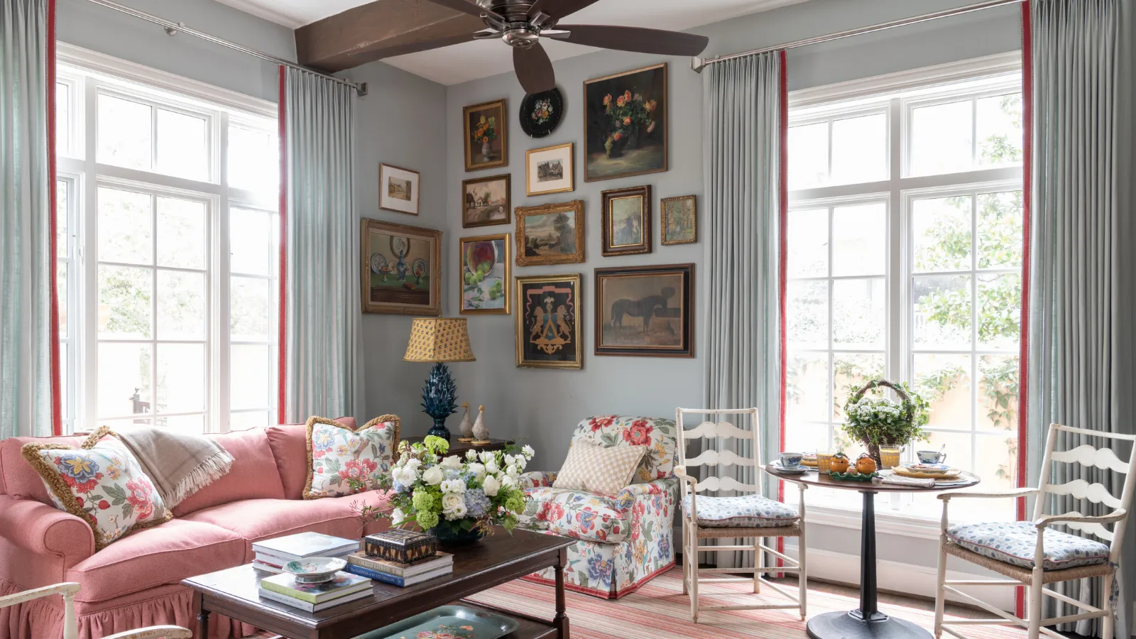 tradičná obývacia izba s veľkým ružovým gaučom, konferenčný stolík s knihami a vázou s kvetmi, kvetinové kreslo a zhluk umenia zavesený v rohu miestnosti' loading='lazy' title=