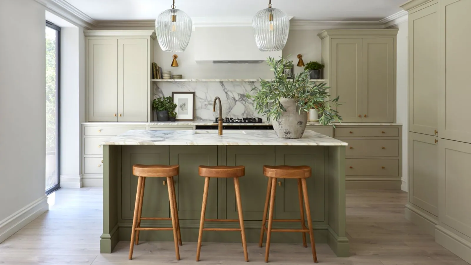 Kuchyňa s šalviam zeleným skrinkou a tmavším zeleným ostrovom. Veľká kamenná váza so zelenými schodmi na ostrove vedľa umývadla a dve sklenené prívesky visiace zo stropu nad.' loading='lazy' title=