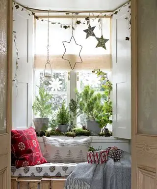 Ideias de decoração de janela de Natal com decorações de estrelas penduradas acima de um assento na janela com almofadas vermelhas e cobertores Scandi
