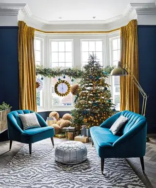 Ideias de decoração de janela de Natal em uma sala de estar azul, com decorações amarelas e azuis em uma guirlanda de folhagens, combinando com a decoração da árvore de Natal