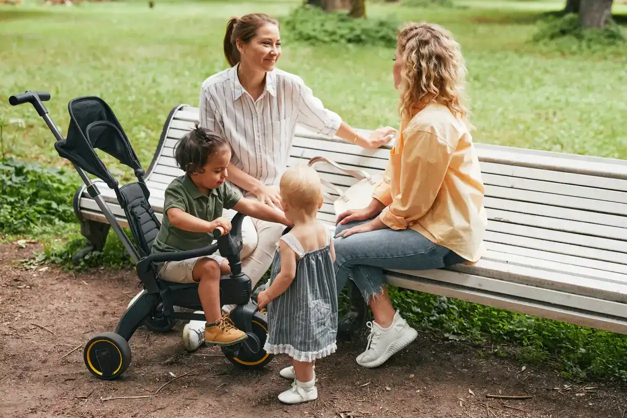 Full length portrait of two young mothers sitting on bench in park and playing with cute babies