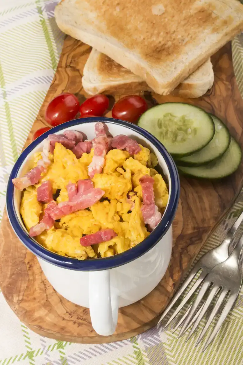 scrambled eggs with roasted bacon in an enamel mug, served with toasted slice of bread, cucumber slices and plum tomatoes on wooden board