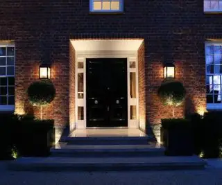 En sort hoveddør med kugleformet buskads på hver side og to væglamper, der oplyser verandaen