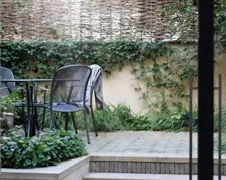 Nahaufnahme eines mehrstöckigen Gartens mit schwarzen Gartenmöbeln, Mauer, Kletterpflanzen und Korbschirm