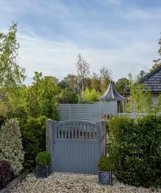 Portão de madeira pintado de cinza rodeado por sebes, com piso de brita.
