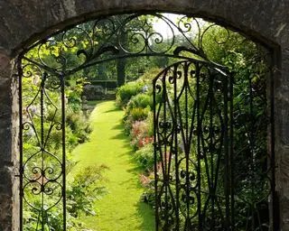 Portão de jardim de ferro forjado ornamentado sob medida em uma parede de pedra, que leva a uma cama formal.