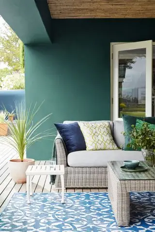 Une terrasse entourée de murs verts avec un tapis à motifs bleus et des meubles en osier