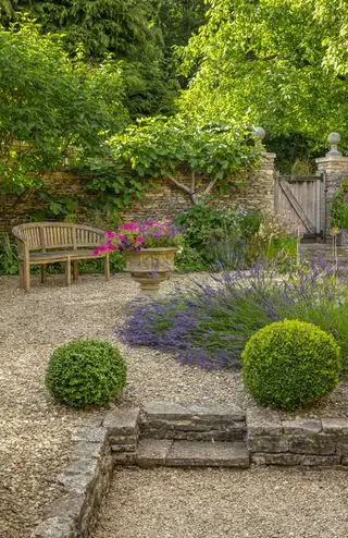 Ideen für Terrassenmöbel im Landhausstil, dargestellt in einer rustikalen Kiesterrasse mit Holzbank, Steinpflanzgefäßen und einem Lavendelblumenbeet.