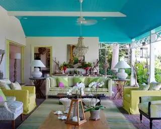 Ampia terrazza all'aperto, con soffitto blu brillante, lampadario con perline, collezione di poltrone colorate, tappeti, cuscini, pareti in mattoni dipinti di bianco, pavimento in mattoni, lampade da tavolo e accessori decorativi e opere d'arte sulla credenza