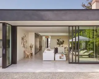 Vista dall'esterno del retro di una proprietà moderna, grandi porte in vetro, che si affacciano sull'ampia zona giorno a pianta aperta, pavimento grigio pietra, tetto nero e soffitto bianco