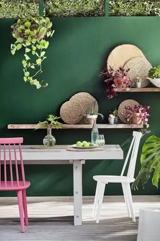 Ideias de decoração de cercas em verde escuro atrás de uma mesa branca com cadeiras pintadas de branco e rosa.