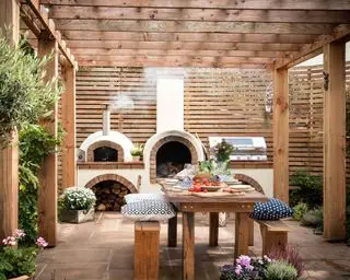 Pergola-Ideen in einem schattigen Bereich mit Holzsitzen, Außenküche und Pizzaofen und abgeschirmtem Zaun.