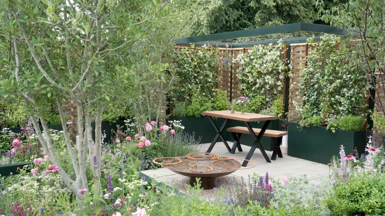 Un giardino moderno con alberi rampicanti fioriti confina con giochi d'acqua e tavoli e sedie da patio all'Hampton Court Flower Show' loading='lazy' title=