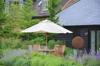 Un coin repas dans la cour avec une table et des chaises en bois et un parasol crème entouré de plantes vertes et violettes